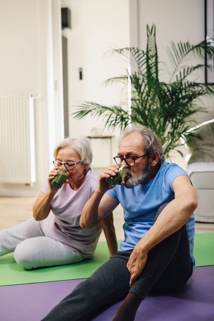 Couple sitting on yoga mats in a bright living room, drinking green smoothies after movement, representing healthy aging, everyday nutrition, and consistent wellness habits.