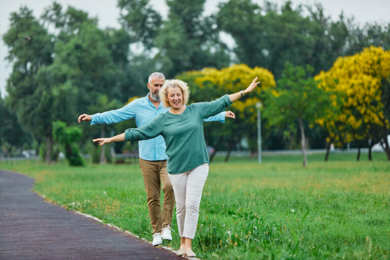 A healthy older couple balance on a curb in a green park.