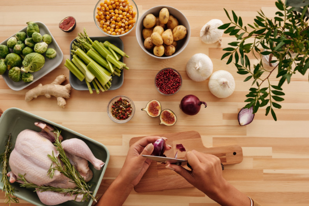 A person engaged in an everyday wellness practice of preparing a meal from whole foods in a warm home kitchen: a whole chicken seasoned with rosemary ready for the oven, and on a butcher-block table, ginger, garlic, crushed red pepper, shallots, figs, Brussels sprouts, dried cranberries, celery, asparagus, potatoes, and chickpeas to complete the meal. 